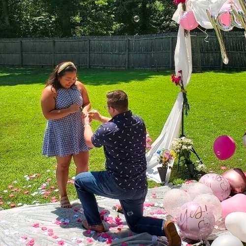 Fall Proposal Ideas: a man on one knee, proposing to his girlfriend in his backyard in front of a pink balloon arch.