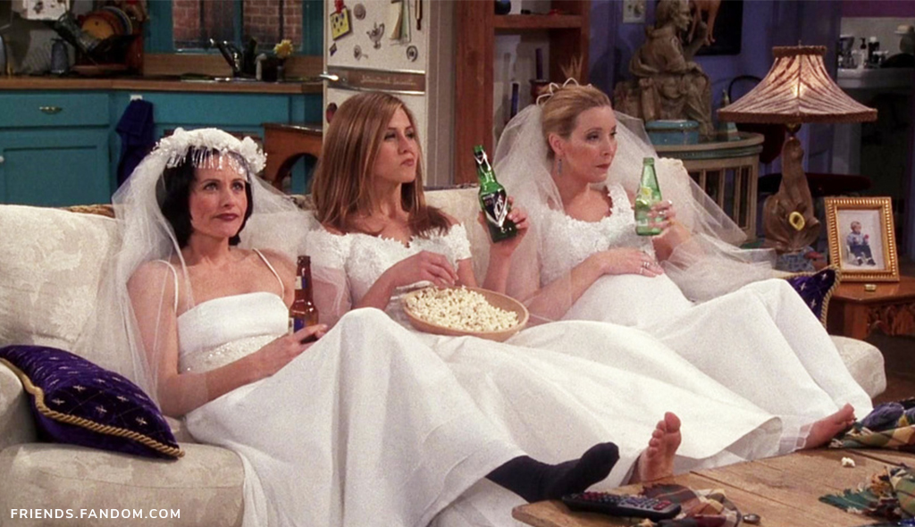 Monica Geller, Rachel Green, and Phoebe Buffay sitting on the couch in wedding dresses drinking beer and eating popcorn.