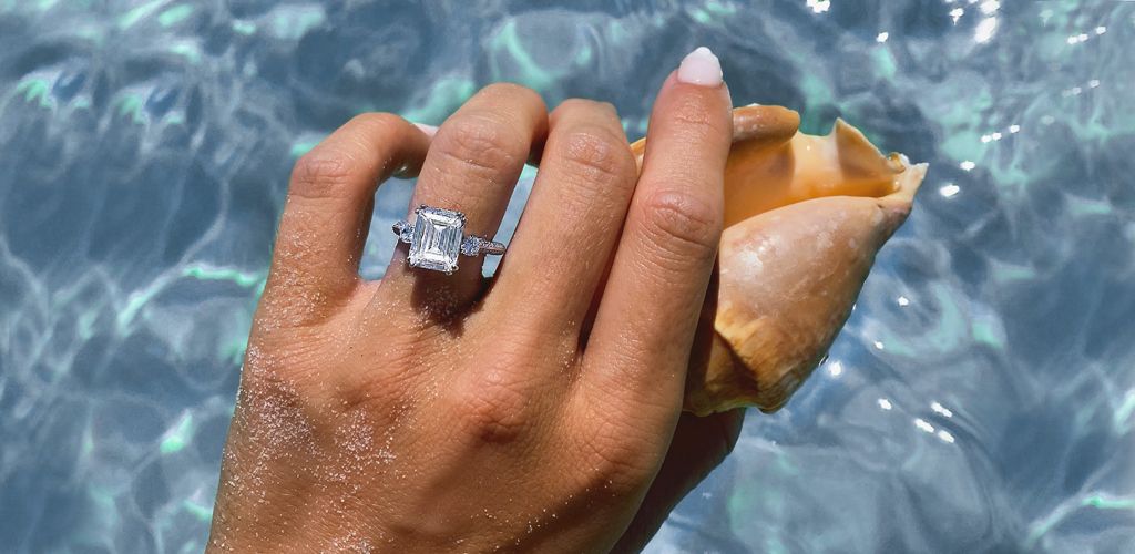 A person holding a sea shell above crystal clear water while showing off her diamond engagement ring. 