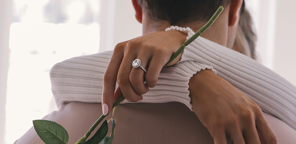 A picture of an engaged couple. The woman has her arms wrapped around his neck and is showing off her diamond ring. 