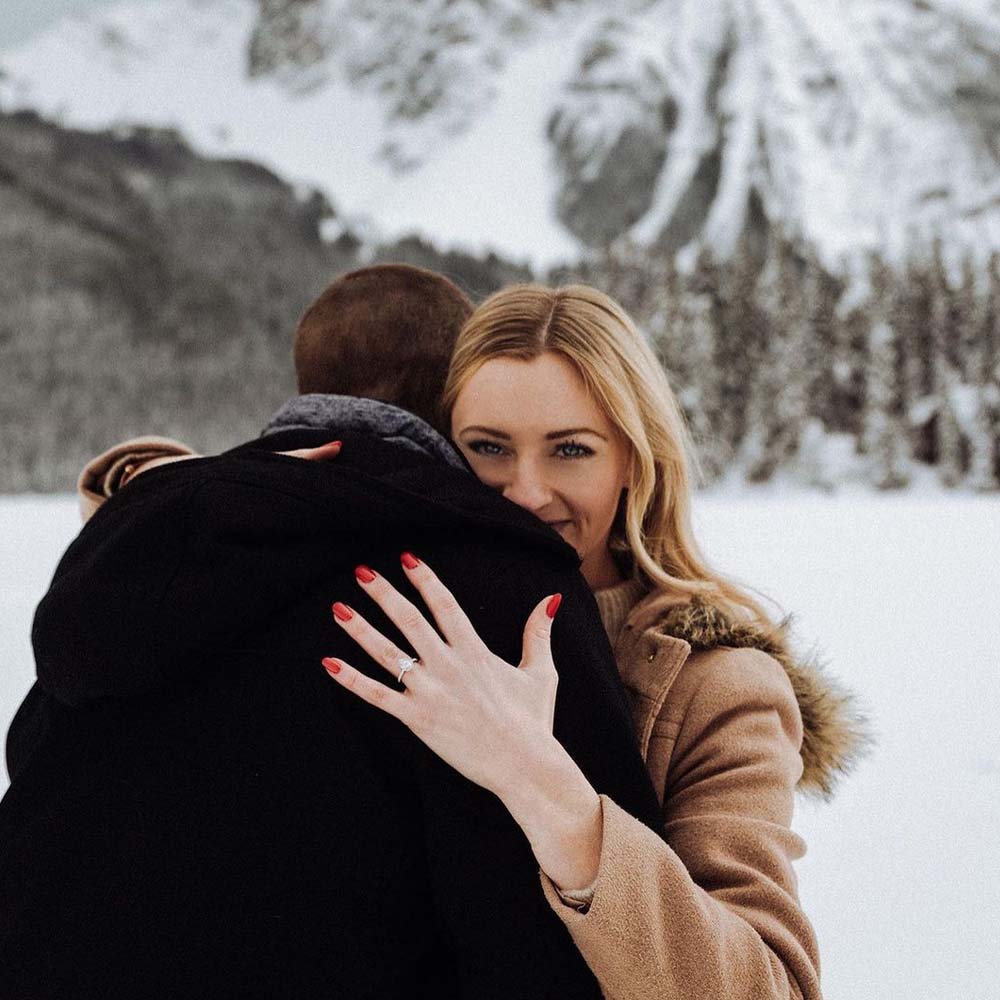 A happy couple embracing in the snow while she shows off her diamond engagement ring 