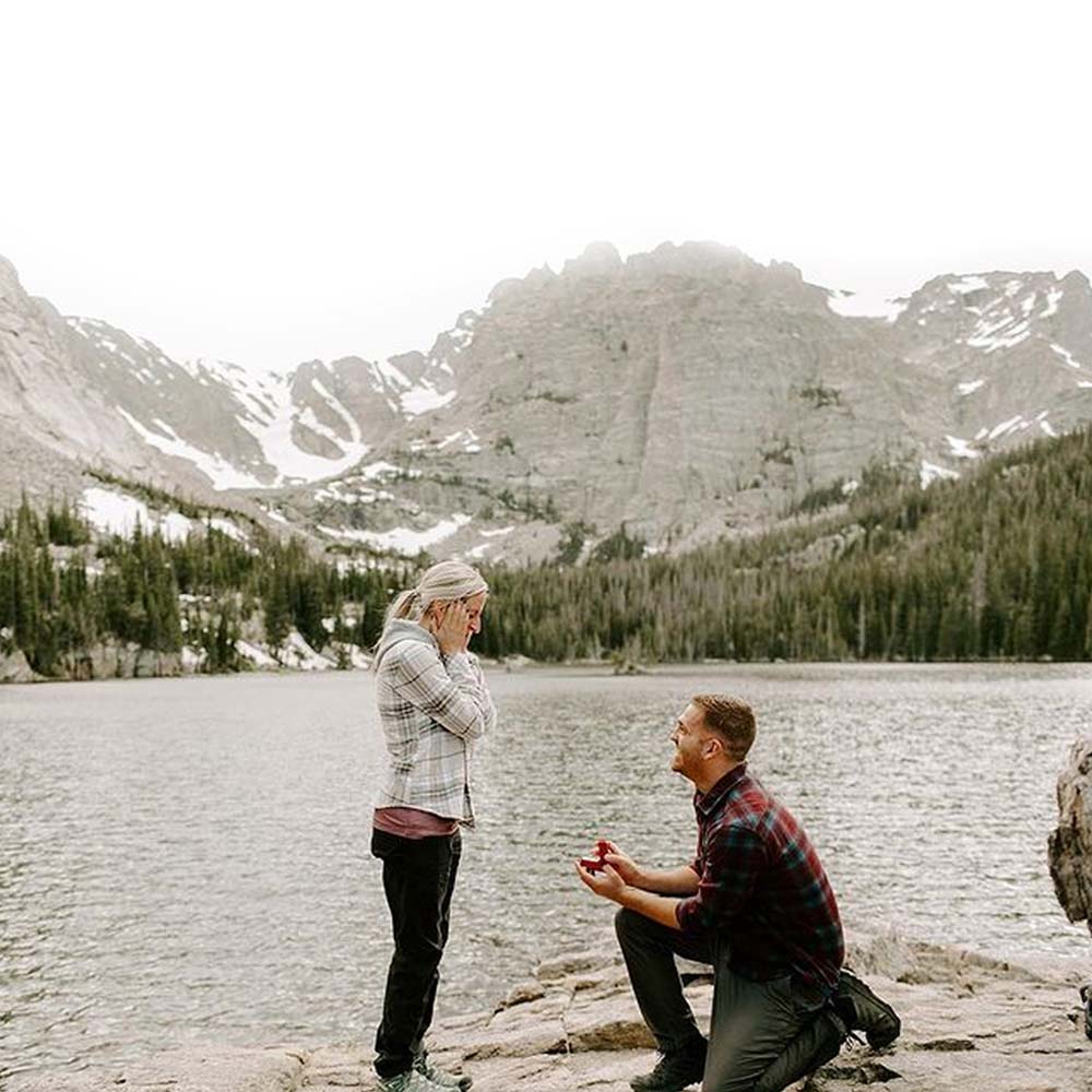 A man proposing to his girlfriend near a lake, forest and mountains 