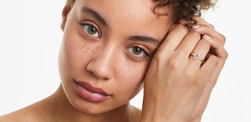 A close-up of a woman touching her face while wearing a gold solitaire ring, used to introduce the look and feel of claw prongs vs. round prongs.