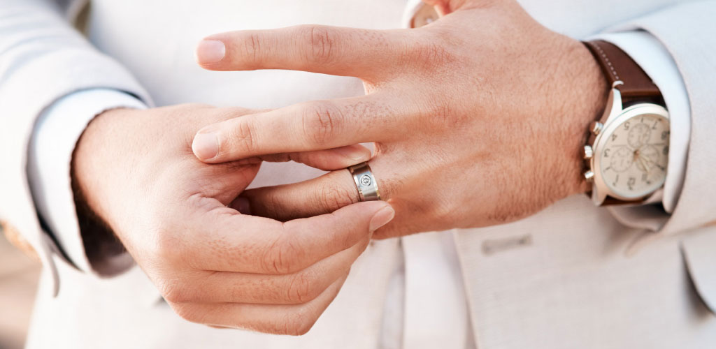 Close-up of a man sliding a band onto his finger while dressed in a light suit, offering a simple example of how men’s engagement rings are worn