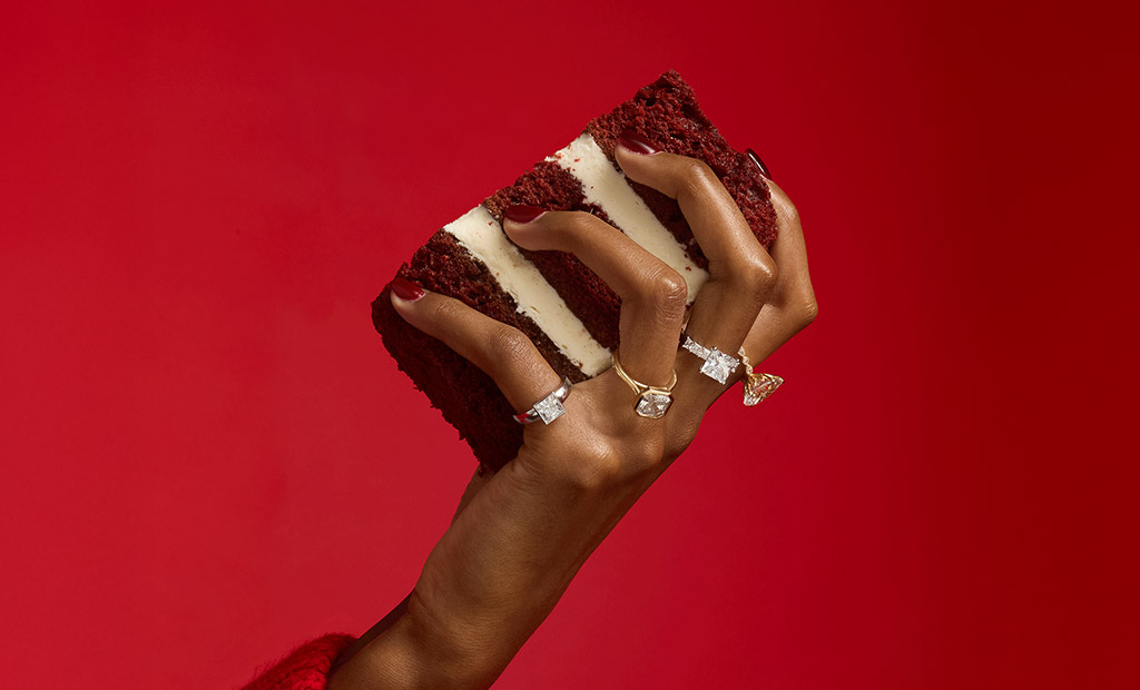 Hand holding a cake slice while wearing diamond rings against a red background for James Allen Valentine’s Day launch.